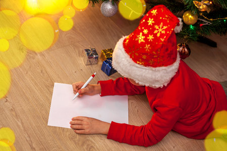 A child in a Santa Claus hat and a red jumper lies on the floor near the Christmas tree and writes a wish list. Holiday concept - Christmas, New Year. Place for textの写真素材