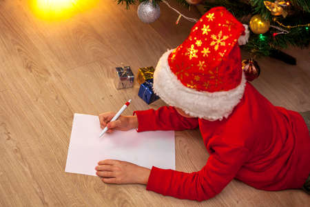 A child in a Santa Claus hat and a red jumper lies on the floor near the Christmas tree and writes a wish list. Holiday concept - Christmas, New Year. Place for text.の写真素材