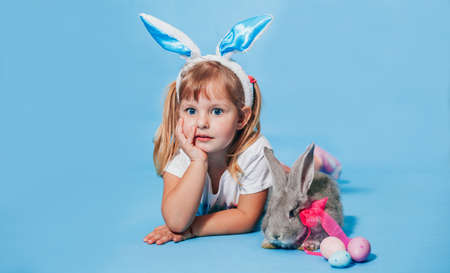 A little pretty girl of European appearance with white hair and with bunny ears on her head sits on a blue background smiling and looking at the camera. Happy easter. Children Protection Dayの写真素材