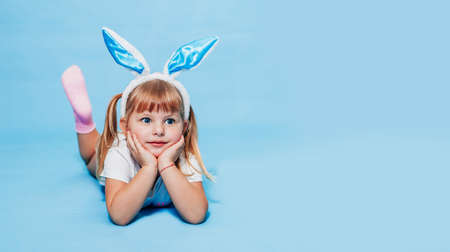 Little blonde girl with bunny ears on her head is lying on a blue background and does not look at the camera. Concept of the holiday - Easter, Christ is resurrected, children's day protection. Bannerの写真素材