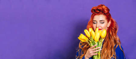Beautiful girl with red hair stands on a purple background and holds a bouquet of yellow tulips. Concept of holiday - March 8, international women's day, Mother's Day. Close-up. Banner. Place for textの写真素材