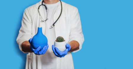 A male doctor holds an enema and a cactus. The concept is the treatment of hemorrhoids, medical care. banner. close-up. Place for text. isolate. medicine. Doctor in a white coat on a blue backgroundの写真素材