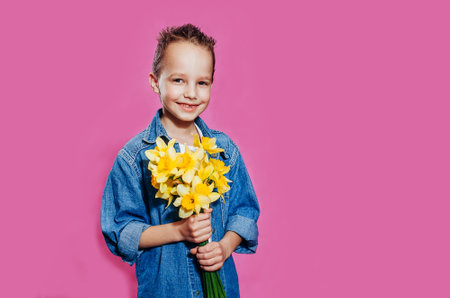 A cute smiling boy looks at the camera and holds a bouquet of yellow narcisses in his hands. A toothless child in a blue shirt stands against a pink background. Mother's Day, March 8.の写真素材
