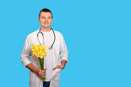 A young doctor men holds a bouquet of yellow narcissists and looks at the camera and stands on a blue background. Concept is Mother's Day, March 8, International Women's Day, Medical Worker's Dayの写真素材