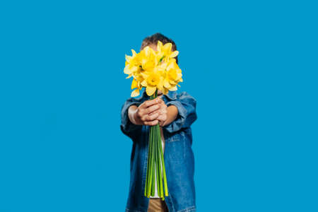A little boy on a blue background holds a bouquet of beautiful yellow daffodils in his hands. Holiday concept - March 8, International Women's Day, Mother's Day. Banner, place for text. Greeting card.の写真素材