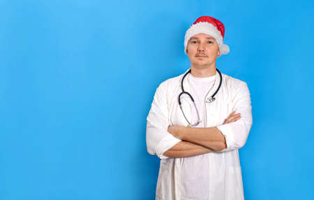 A young doctor in a red New Year's hat of Santa Claus and a white coat looks into the camera and smiles. Doctor Santa Claus on a blue background. Concept - new year, Christmas, congratulationsの写真素材