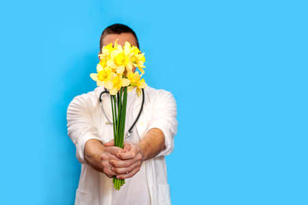 A young doctor men holds a bouquet of yellow narcissists and stands on a blue background. Concept is Mother's Day, March 8, International Women's Day, Medical Worker's Day. Daffodils close up.の写真素材