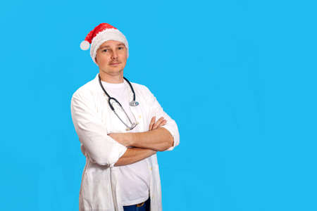A young doctor in a red New Year's hat of Santa Claus and a white coat looks into the camera and smiles. Doctor Santa Claus on a blue background. Concept - new year, Christmas, congratulations.の写真素材