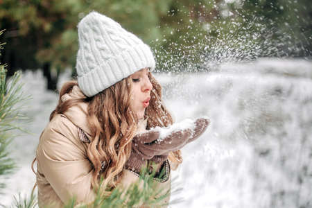 Beautiful blonde girl close-up in a white winter hat against the backdrop of a snowy winter forest. A pretty girl blows snow from her palms. Place for text, banner. Christmas, new year.の写真素材