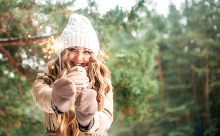 A beautiful blonde girl in a white winter hat and a beige coat holds sparklers in her hands and looks into the camera and smiling. Place for text, banner, new year, Christmasの写真素材