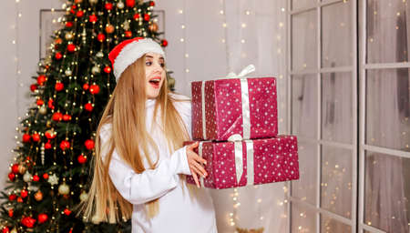 A beautiful girl in a Santa Claus hat holds gifts and smiles. Blonde near the Christmas tree. Christmas. New Year. A place for text.の写真素材