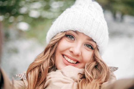 Young beautiful happy girl in a white winter knitted hat makes a selfie in the winter forest. Emotional curly woman smiles to the camera. Close-up. Walk through a frosty pine groveの写真素材