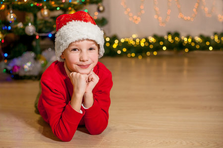 A child in Santa Claus' red hat lies near the Christmas tree and looks into the camera. Space for text. Christmas, New Year's Day, St. Nicholas Day. Bokeh of garlandsの写真素材