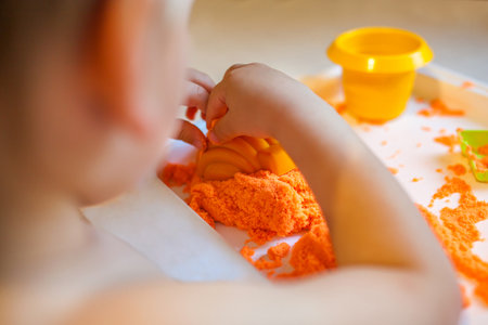Little child playing with orange kinetic sand at white wooden table, selective focus. Smiling happy child. Hands close upの写真素材