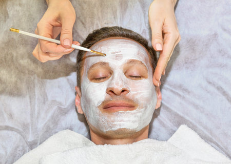 Face of laying man with cream mask, hands of beautician with brush, beauty, spa