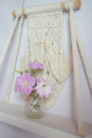 Wall decor in ecological boho, Scandinavian style. Wooden Shelf with petunia flowers in the macrame technique. Interior decor close-up. Macrame decor on a light background. Vertical orientationの写真素材