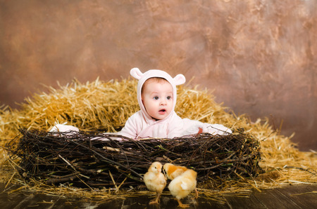 little girl. child in pink overalls with ears.Sitting in a nest made of twigs on the background of hay with chicksの写真素材