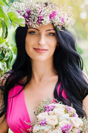 Beautiful brunette girl in a wreath of flowers, holding in his hand bouquet. Portrait of bride.の写真素材