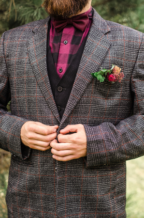 The young man wears a gray jacket and buttoning.Cool Boutonniere on hip trendy groom at wedding. Grey suit, colorful vinous boutonniere. Maroon Bow tie.の写真素材
