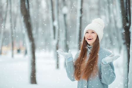 Outdoor close-up portrait of young beautiful happy smiling girl holding snow in hands. Dressed in winter clothes and a white hat, standing in the winter forest.の写真素材