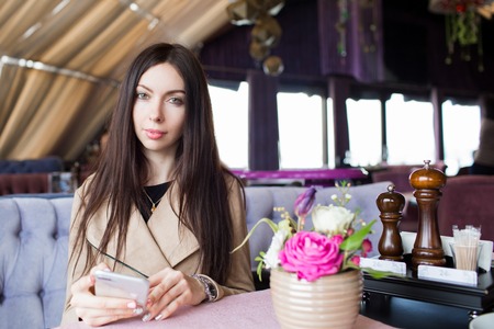 Close-up, sitting in a cafe-restaurant, waiting for her order. Beautiful brunette with phone in hands. The woman at the table.の写真素材