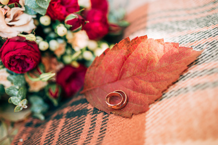 Wedding rings on autumn leaf. Accessories for the autumn wedding. Beautiful wedding bouquet of red and beige roses.の写真素材