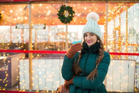 Beautiful smiling girl holding a cup of tea or coffee in winter. Beautiful woman in the winter on the street with New Year decorations. Christmas Fair.の写真素材