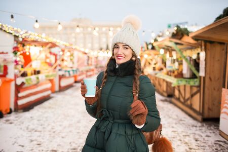 Beautiful smiling girl holding a cup of tea or coffee in winter. Beautiful woman in the winter on the street with New Year decorations. Christmas Fair.の写真素材