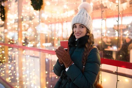 Beautiful smiling girl holding a cup of tea or coffee in winter. Beautiful woman in the winter on the street with New Year decorations. Christmas Fair.の写真素材