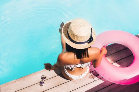 Beautiful and young mulatto near the pool, takes sun baths and sunbathes. Rear view of sexy woman in bikini sitting on the edge of pool. Summer vacation. Girl in a fat hat and glasses.の写真素材