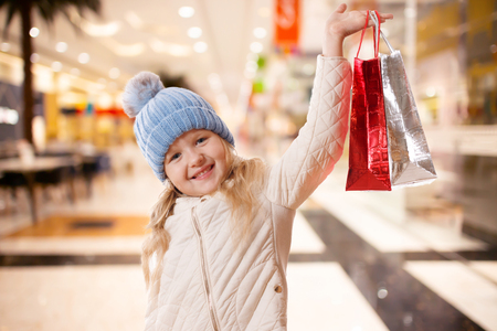 A little girl in a blue hat with a pompon and a beige jacket with paper bags. The concept of purchases and sales. In the mallの写真素材