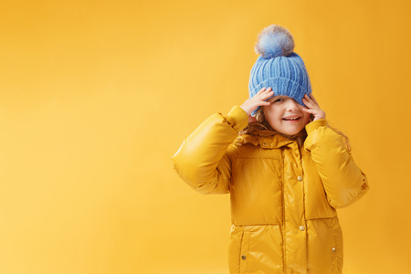 Portrait happy little girl preschooler in a winter jacket and blue hat. Yellow on a yellow background.の写真素材