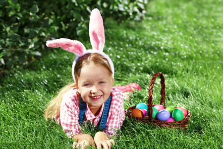A cheerful little girl preschooler dressed in bunny ears is lying on the lawn with a basket of painted Easter eggs. Happy Easterの写真素材