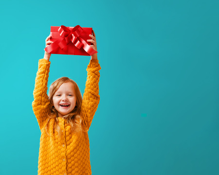 Portrait of a little girl in a warm mustard knitted sweater on a cyan background. A child holds a red box with a gift above his head. The concept of celebration, giving and receiving a gift.の写真素材