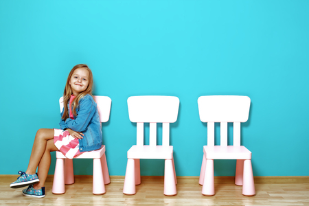 Happy little child  girl is sitting on the chair against the background of the blue wall. Nearby are empty chairs. The concept of happiness, childhood, peopleの写真素材
