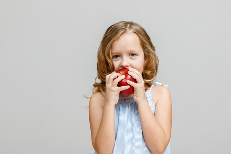 Portrait of a happy smiling little blonde girl on a gray background. Baby eating red appleの写真素材