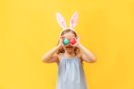Cheerful little child girl with bunny ears with two Easter eggs on a colored background.の写真素材