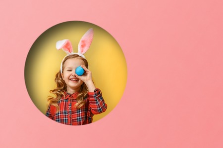 Cheerful little girl with bunny ears with an easter egg. Kid in a round hole circle in colored yellow and pink backgroundsの写真素材