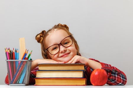 Cute little girl wearing glasses is sitting at the table, leaning on thick books. Education conceptの写真素材