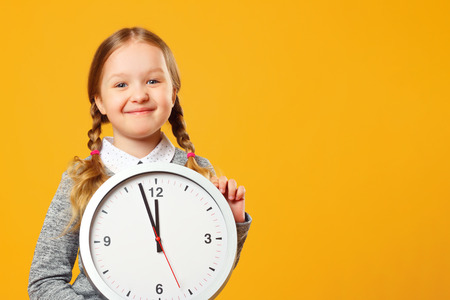 Little girl holding a big clock on a yellow background. The concept of education, school, deadline, time to learn.の写真素材