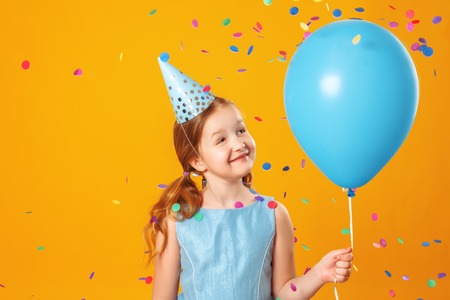 Cheerful little girl at a party. A child holds a blue ball in a rain of confetti. Closeup portrait on yellow background.の写真素材