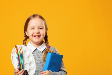 Portrait of a cheerful little girl on a yellow background. Schoolgirl holds a book, pencils, brushes and a palette. The concept of education. Copy space.の写真素材