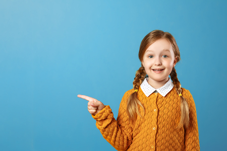Closeup portrait of a little girl schoolgirl on a blue background. The child points to the side. Copy space.の写真素材