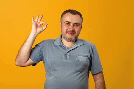 A young man in a gray polo shirt shows the ok sign and looks into the camera. Yellow background Close-up.の写真素材