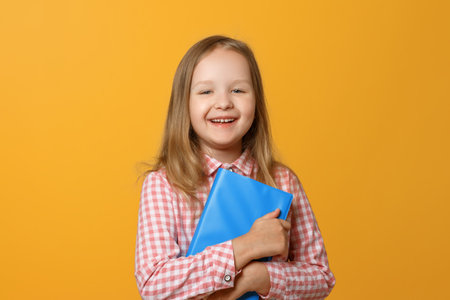 Portrait of a merry little blond girl on a yellow background. A child in a pink shirt and book is...