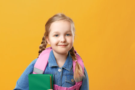 Portrait of a little girl schoolgirl with a backpack and book on a yellow background. Baby closeup....