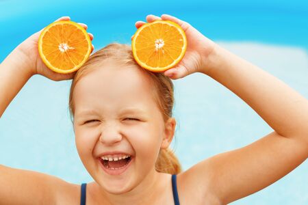 Funny little girl swimming in the pool. The child holds the halves of the orange instead of the ears. Summer concept.の写真素材