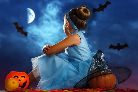 Halloween celebration. A little girl dressed as Princess Cinderella sits with her back surrounded by pumpkins and looks at the evening moonlit sky.の写真素材