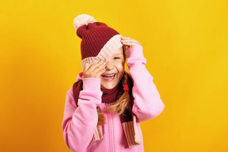 Portrait of a cheerful little girl in a scarf and sweatshirt on a yellow background. The child peeks out from under the hat.の写真素材
