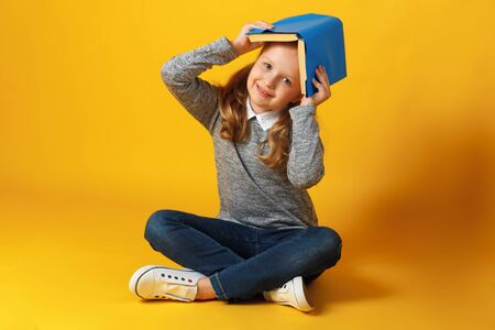A cheerful little student girl is sitting on the floor with a book on her head. Education and school concept.の写真素材